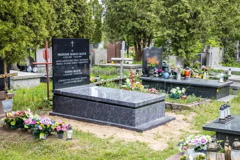 Kolorowe zdjęcie: nowy granitowy nagrobek Mariana Mazura wśród drzew i sąsiednich grobów, przed nim sztuczne kwiaty.