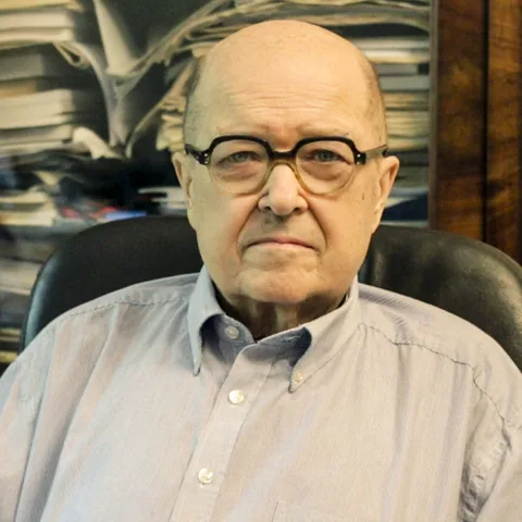 Kolorowe zdjęcie: Józef Kossecki w okularach i jasnej koszuli, w fotelu, w tle regał z papierami.